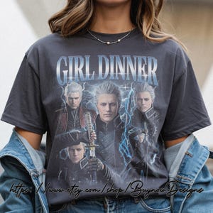 May include: Dark gray t-shirt featuring the text "GIRL DINNER" above a graphic of a character with white hair and a sword. The graphic has a blue lightning effect. The shirt is a relaxed fit.