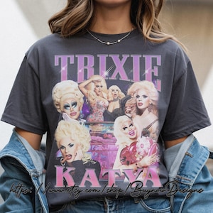 May include: Dark gray t-shirt featuring a collage of images of Trixie and Katya, with the names in pink block letters. The shirt has a vintage, distressed look. The Etsy shop URL is visible at the bottom.