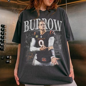 Op de afbeelding: Een antracietgrijs t-shirt met korte mouwen en een grote grafische print van een footballspeler in een zwart shirt met het nummer 9. De tekst BURROW staat in grote witte letters boven de speler en een gouden handtekening is zichtbaar.