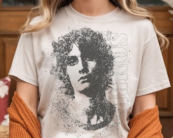 May include: Beige t-shirt featuring a grayscale portrait of a person with curly hair and the word "SOMBRD" printed vertically on the right side. The portrait has a distressed, speckled effect.