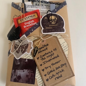 May include: A brown paper package wrapped with twine and decorated with stickers, including a skull and crossbones, a bat, and a tea bag. The package is labeled with the text "Opposites Attract Creatures of the Night Illustrated Dry Humor Gothic Love Story Cute, quick read".