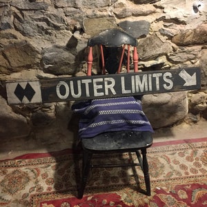 May include: A black wooden chair with a blue and white striped blanket on the seat. The chair is in front of a stone wall with a wooden sign that reads "OUTER LIMITS" with a black arrow pointing to the right and a black chevron pointing to the left. The chair is on a red and cream patterned rug.
