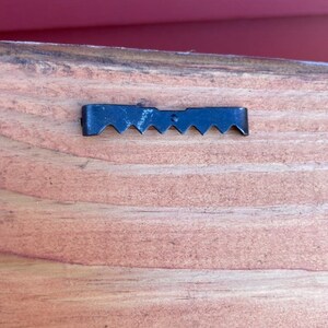May include: A close-up of a black metal sawtooth hanger attached to the back of a wooden sign.