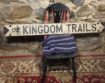 May include: A white wooden sign with black lettering that reads "KINGDOM TRAILS". A black silhouette of a person riding a bicycle is on the left side of the sign. The sign is hanging on a stone wall.