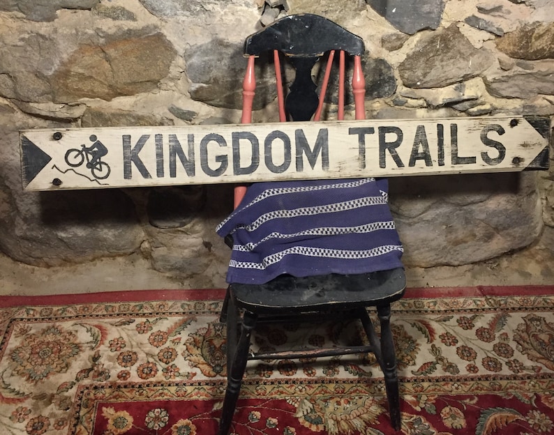 May include: A white wooden sign with black lettering that reads "KINGDOM TRAILS". A black silhouette of a person riding a bicycle is on the left side of the sign. The sign is hanging on a stone wall.