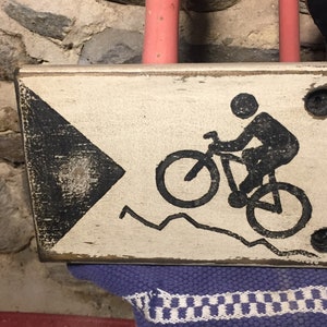 May include: A white wooden sign with black painted details. The sign features a black silhouette of a person riding a bicycle on a mountain path. The sign has a black arrow pointing to the right.