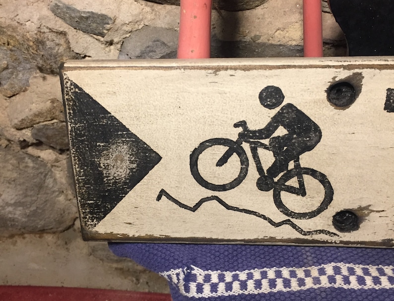 May include: A white wooden sign with black painted details. The sign features a black silhouette of a person riding a bicycle on a mountain path. The sign has a black arrow pointing to the right.