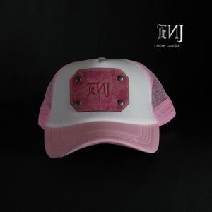 May include: A light pink and white trucker hat featuring a rectangular pink leather patch on the front panel. The patch is secured with four silver-toned metal rivets and displays the embossed text ENJ in a stylized gothic font.