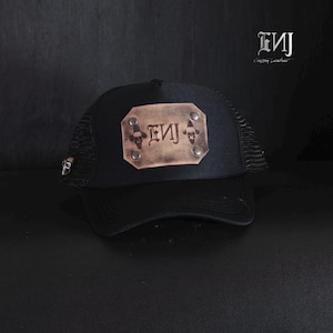 Puede incluir: Una gorra de camionero negra con un parche rectangular de cuero color canela en el panel frontal. El parche está asegurado con cuatro remaches plateados y muestra el texto ENJ con dos pequeños iconos de calaveras. Una pequeña calavera de metal está en la malla.