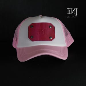 May include: A white and light pink trucker hat featuring a rectangular magenta leather patch on the front panel. The patch is secured with four silver-toned metal rivets and displays the embossed text ENJ. The hat has a mesh back and sides.