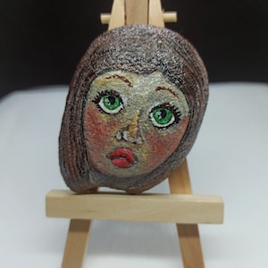 Puede incluir: Una piedra pintada con una cara. La piedra está pintada para parecer una persona con cabello castaño, ojos verdes y labios rojos. La piedra está en un pequeño caballete de madera.