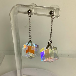 Op de afbeelding: Een paar zilverkleurige oorhangers. Elke oorbel heeft een kleine, iriserende kubusvormige hanger die aan een ketting hangt. De kubussen reflecteren meerdere kleuren, met een zilveren bol aan de bovenkant.