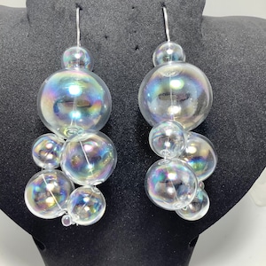 May include: A pair of iridescent bubble earrings with a silver hoop and multiple bubble-shaped charms. The earrings are made of glass and have a rainbow iridescent finish.