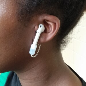 May include: White wireless earbud with a silver chain attached, worn in the ear. The earbud is connected to a small, clear silicone tip. The person is wearing a black shirt.