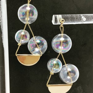 Puede incluir: Un par de pendientes de oro con cuentas de vidrio iridiscente en forma de burbuja. Los pendientes están suspendidos de un alambre de oro y tienen una forma de media luna de oro en la parte inferior.