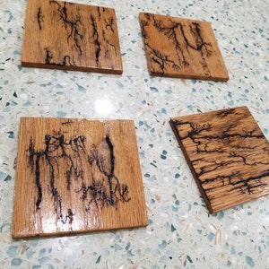 Puede incluir: Cuatro posavasos cuadrados de madera con un acabado de madera natural y patrones de rayos negros. Los posavasos están sobre una superficie blanca con pequeñas motas azules y blancas.