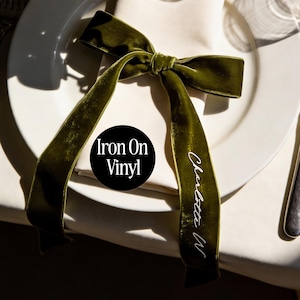 May include: An olive green velvet ribbon bow on a white plate. The ribbon is inscribed with the name "Charlotte W" in white script. A black circle with the text "Iron On Vinyl" is also visible.