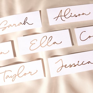 May include: A set of ten white place cards with gold foil lettering. The names on the cards are Sarah, Alison, Ella, Court, Ian, Taylor, and Jessica.