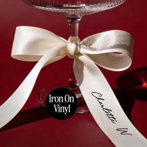 May include: A clear glass stemware piece featuring a tied cream-coloured satin ribbon around its stem. The ribbon displays the black printed text "Charlotte W" and a circular black label with the white text "Iron On Vinyl" is visible.