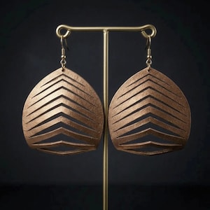 May include: A pair of copper-colored, teardrop-shaped earrings with a layered chevron design. The earrings are suspended from gold-colored ear wires and are displayed on a gold-colored earring stand against a dark background.
