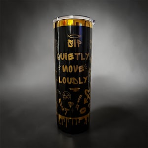 May include: A tall, cylindrical black tumbler with a metallic gold rim and base. The body features gold text that reads "SIP QUIETLY MOVE LOUDLY" along with various small gold graphic icons including a diamond, a lightning bolt, and a heart.