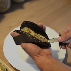 Puede incluir: Una persona está usando un cuchillo para ostras para abrir una ostra que se sostiene en un guante de cuero marrón para ostras. La ostra está en un plato blanco.