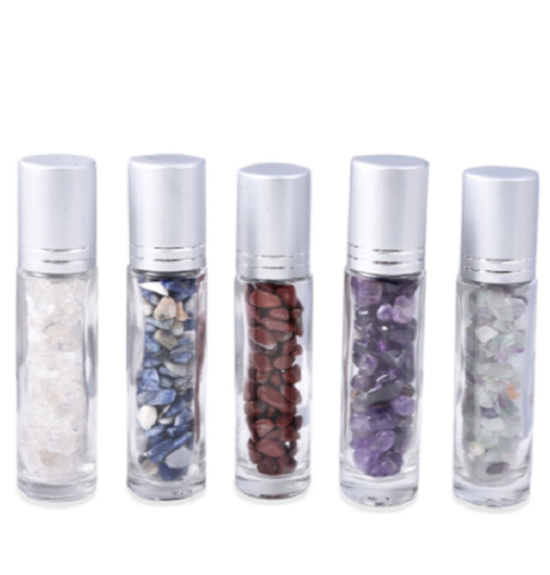 May include: Five clear glass roller bottles with silver caps filled with different colored stones. The bottles are arranged in a row from left to right. The stones are white, blue, red, purple, and green.
