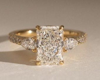 May include: A gold engagement ring with a large, rectangular-cut diamond as the centerpiece. Two smaller pear-shaped diamonds flank the main stone. The band is also adorned with smaller accent stones, creating a sparkling effect.