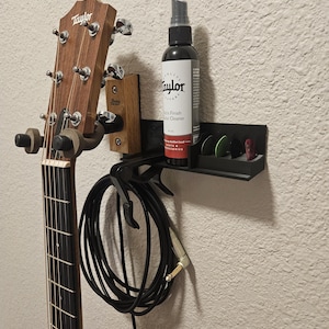 May include: A black wall-mounted guitar stand with a Taylor acoustic guitar hanging on it. The stand has a built-in holder for guitar picks and a bottle of Taylor Guitar Finish and Guitar Cleaner. A black guitar cable is hanging from the stand.