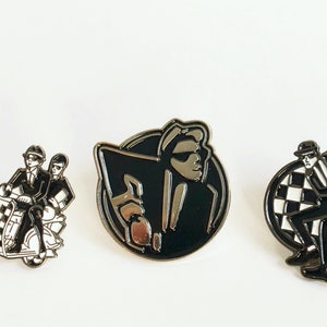 May include: Three black and white enamel pins featuring a band. One pin shows two figures riding a scooter, another shows a single figure in a suit, and the last pin shows a figure in a suit with a checkerboard background.
