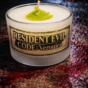 Könnte beinhalten: Eine runde, mattierte Glas-Kerze mit dem Text "RESIDENT EVIL CODE: Veronica" auf einem Etikett. Die Kerze hat weißes Wachs und ein grünes Blatt oben. Die Oberfläche hat ein dunkles, strukturiertes Aussehen mit roter Flüssigkeit.