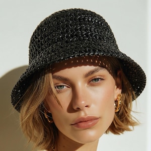 May include: A black woven bucket hat with a rounded crown and a downward-sloping brim. The hat is worn with a cream-coloured sweater with navy blue trim and a gold chain necklace. The model has short blonde hair and gold hoop earrings.