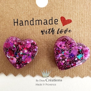 May include: A pair of pink heart-shaped earrings with glitter embedded in the resin. The earrings are on a brown card with the text "Handmade with love" and "By Dom Creations Made in Provence".