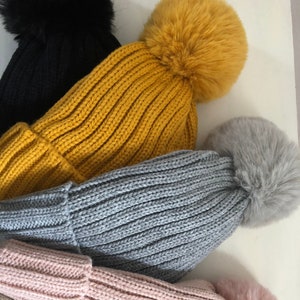 Può includere: Cinque berretti di lana con pon pon in pelliccia sintetica di vari colori, tra cui nero, giallo, grigio, rosa e crema.