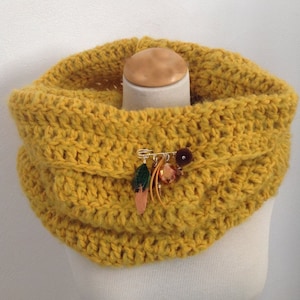 May include: Mustard yellow crocheted cowl neck warmer with a decorative pin. The pin features a small green leaf, a copper-colored feather, and a cluster of amber-colored beads. The cowl is displayed on a mannequin.
