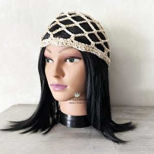 May include: A beige crochet head covering with a net-like design. The head covering is worn on a mannequin with black hair and a black choker. The head covering is made of a light-colored yarn.
