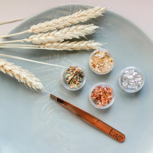 May include: Four small containers of gold, silver, iridescent, and copper leaf flakes on a light blue plate with a pair of tweezers labeled "Love". A bundle of dried wheat stalks is in the background.