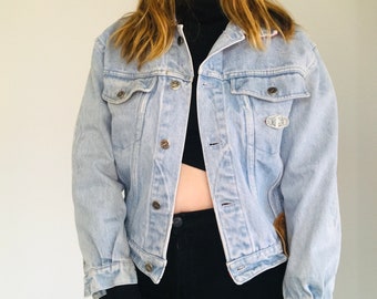 May include: Light blue denim jacket with silver-tone buttons and a small decorative pin. The jacket has two chest pockets and a classic collar. The sleeves are long, and the jacket appears to be slightly oversized.