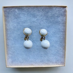 May include: A pair of white, faceted ball earrings with gold-coloured accents. The earrings are displayed in a beige box with white padding. The earrings have a classic, elegant design.