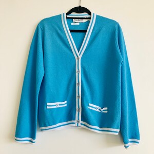 May include: A light blue cardigan sweater with white and light blue striped trim on the collar, pockets, and cuffs. The sweater has a V-neck, button closure, and two front pockets. The brand label "TALBOTT" is visible.