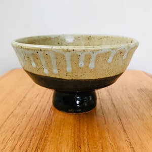 May include: A handmade ceramic pedestal bowl with a speckled beige exterior and a dark brown base. The rim features white glaze drips. The bowl is set on a wooden surface.