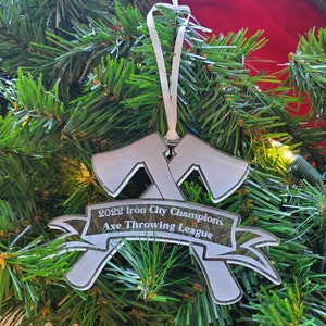 May include: A white acrylic Christmas ornament shaped like two crossed axes with a banner that reads "2022 Iron City Champions Axe Throwing League".