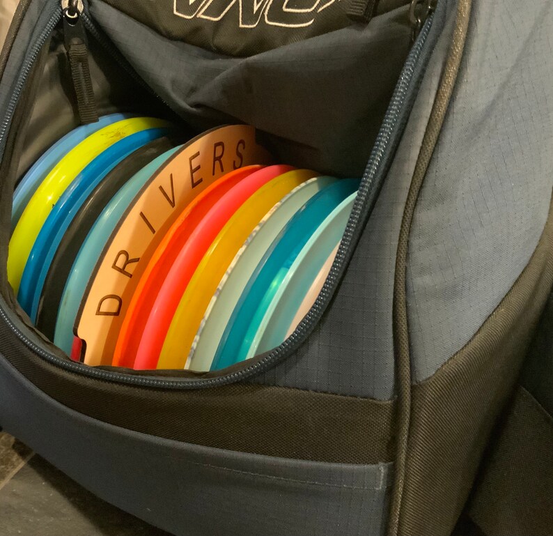 Op de afbeelding: Een blauwe discgolf-tas met een ritsvak dat open is en gevuld is met een stapel kleurrijke discgolf-schijven. De schijven zijn gelabeld "DRIVERS" op een houten bord.