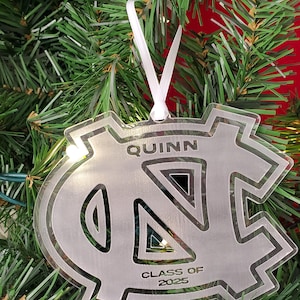 May include: Clear acrylic ornament in the shape of a stylized letter "N" and "C" with the name "QUINN" and "CLASS OF 2025" etched into the surface. The ornament hangs from a white ribbon against a backdrop of a Christmas tree.
