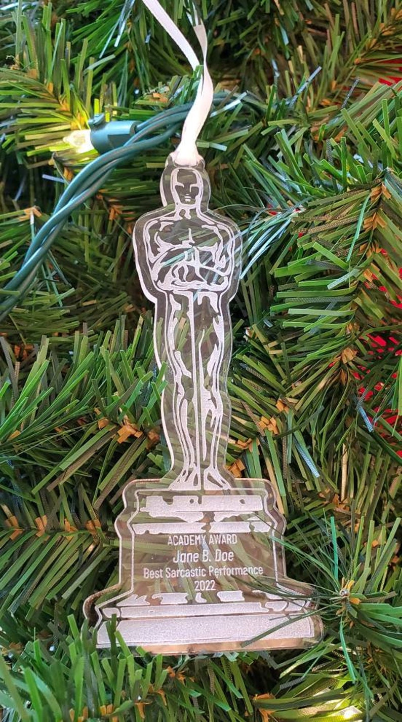 Puede incluir: Un adorno de pl&aacute;stico transparente con forma de estatuilla de los Oscar. El adorno tiene el texto "ACADEMY AWARD" y "Jane B. Doe" en la base. El texto "Best Sarcastic Performance 2022" tambi&eacute;n est&aacute; en la base.