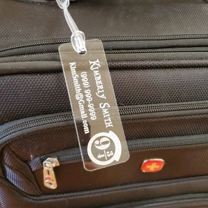 May include: A black luggage tag with the name Kimberly Smith, a phone number (989) 999-9999, and an email address KimSmith@Gmail.com. The tag is attached to a black suitcase with a red Swiss cross logo.