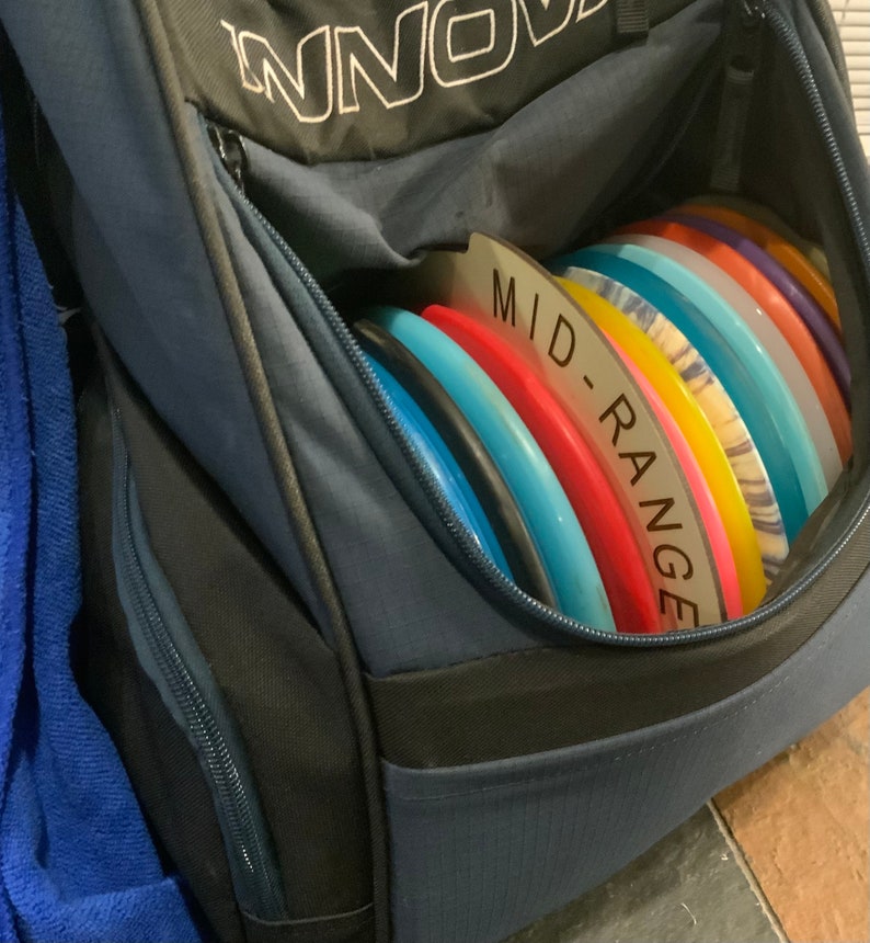Op de afbeelding: Een blauwe discgolf-tas met een ritsvak en het woord "INNO" gedrukt op de voorkant. De tas is gevuld met een stapel kleurrijke discgolf-schijven, met het label "MID-RANGE".