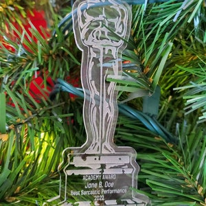 Puede incluir: Un adorno de pl&aacute;stico transparente con forma de la estatuilla del Premio de la Academia. La base del adorno tiene el texto "ACADEMY AWARD Jane B. Doe Best Sarcastic Performance 2022".