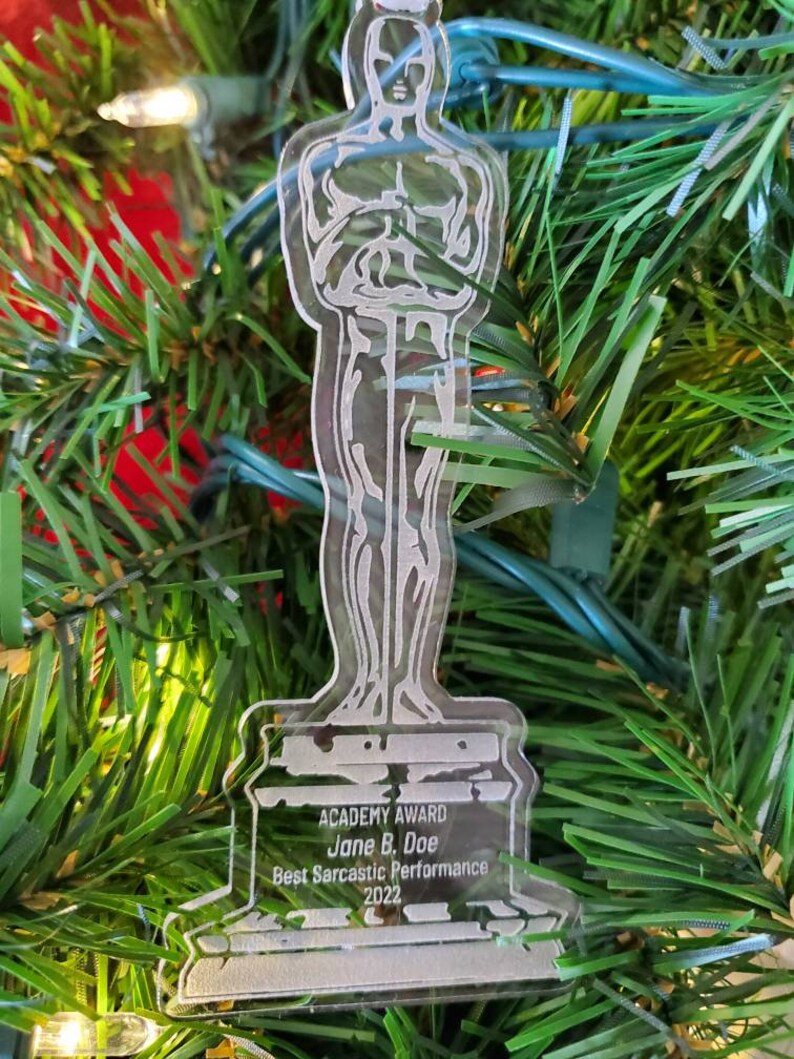 Puede incluir: Un adorno de pl&aacute;stico transparente con forma de la estatuilla del Premio de la Academia. La base del adorno tiene el texto "ACADEMY AWARD Jane B. Doe Best Sarcastic Performance 2022".