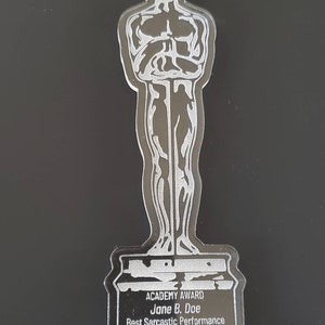 Puede incluir: Una estatuilla de Oscar de pl&aacute;stico plateado con una cinta blanca. La base de la estatuilla dice "ACADEMY AWARD Jane B. Doe Best Sarcastic Performance 2022".